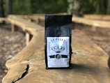 A bag of coffee labeled 'EPIPHANY' with a logo and details including 'Whole Bean' and 'Medium Roast' is placed on a long wooden table outdoors. The background is blurred, showing a natural setting with trees and more wooden tables.