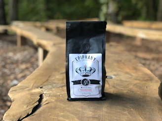 A bag of coffee labeled 'EPIPHANY' with a logo and details including 'Whole Bean' and 'Medium Roast' is placed on a long wooden table outdoors. The background is blurred, showing a natural setting with trees and more wooden tables.