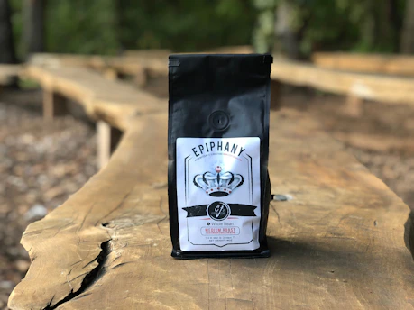 A bag of coffee labeled 'EPIPHANY' with a logo and details including 'Whole Bean' and 'Medium Roast' is placed on a long wooden table outdoors. The background is blurred, showing a natural setting with trees and more wooden tables.