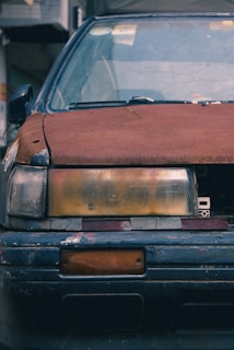 A close-up view of the front of an old, weathered car with a rusted hood and faded, cracked headlight covers. The paint is chipped with visible signs of wear and tear, and the license plate area is empty.