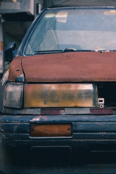 A close-up view of the front of an old, weathered car with a rusted hood and faded, cracked headlight covers. The paint is chipped with visible signs of wear and tear, and the license plate area is empty.