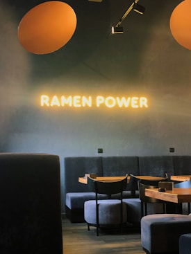 A cozy interior of a restaurant with gray upholstered seating and wooden tables. The wall displays a neon sign that reads 'RAMEN POWER,' creating a warm atmosphere. Overhead lighting adds to the soft, inviting ambiance.