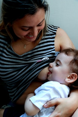 A woman is breastfeeding a baby while gazing at them with a smile. The woman is wearing a striped shirt, and there is a sense of warmth and care in her expression. The baby, dressed in a white shirt, is nestled comfortably in her arms.