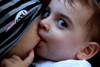 A close-up of a baby breastfeeding, with the child looking intently to the side. The baby has bright blue eyes and soft, light brown hair. The mother is wearing a striped garment with a small heart and the word 'cool'.