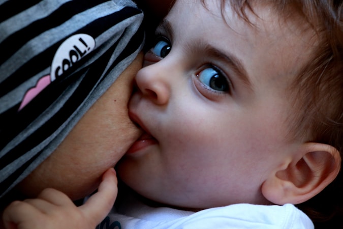 A close-up of a baby breastfeeding, with the child looking intently to the side. The baby has bright blue eyes and soft, light brown hair. The mother is wearing a striped garment with a small heart and the word 'cool'.