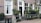 A charming brick building facade with large windows adorned with white curtains. An array of potted plants is arranged on a ledge by the entrance, creating a lush, green atmosphere. The street sign identifies the location as Herenmarkt Centrum. A classic street lamp is mounted above the entrance.
