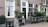 A charming brick building facade with large windows adorned with white curtains. An array of potted plants is arranged on a ledge by the entrance, creating a lush, green atmosphere. The street sign identifies the location as Herenmarkt Centrum. A classic street lamp is mounted above the entrance.