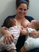 A woman is smiling while breastfeeding two babies. The babies are wearing matching floral outfits, and the woman appears relaxed and content.