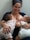 A woman is smiling while breastfeeding two babies. The babies are wearing matching floral outfits, and the woman appears relaxed and content.