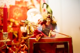 A whimsical puppet with a green hat and curly blonde hair sits on display holding a thin wand or stick. Surrounding the puppet are bright red decorations, including a candle and various boxes, creating a festive and magical atmosphere.
