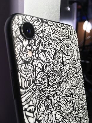A smartphone with a black and white doodle-like pattern on its back cover, featuring intricate and abstract designs. The phone's camera lens is prominently visible. The background is blurred with subtle lighting.