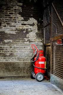 A red fire extinguisher with wheels is positioned against a worn, textured brick wall, surrounded by a metal fence. The setting appears industrial and slightly neglected.