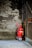 A red fire extinguisher with wheels is positioned against a worn, textured brick wall, surrounded by a metal fence. The setting appears industrial and slightly neglected.