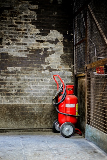 A red fire extinguisher with wheels is positioned against a worn, textured brick wall, surrounded by a metal fence. The setting appears industrial and slightly neglected.