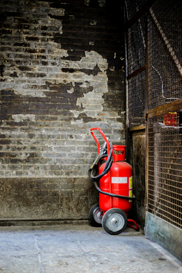 A red fire extinguisher with wheels is positioned against a worn, textured brick wall, surrounded by a metal fence. The setting appears industrial and slightly neglected.