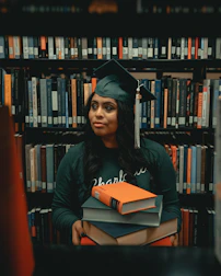 A graduation cap resting on a stack of books with a cross necklace beside it.