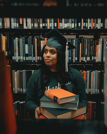 A person wearing a graduation cap and gown is holding a stack of books. They are standing in front of bookshelves filled with various books in a library setting. The person has a calm and thoughtful expression, surrounded by a plethora of literature.