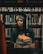 A person wearing a graduation cap and gown is holding a stack of books. They are standing in front of bookshelves filled with various books in a library setting. The person has a calm and thoughtful expression, surrounded by a plethora of literature.