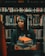 A person wearing a graduation cap and gown is holding a stack of books. They are standing in front of bookshelves filled with various books in a library setting. The person has a calm and thoughtful expression, surrounded by a plethora of literature.