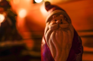 A small Santa Claus figurine with a long white beard and red attire stands out against a warm, blurred background. The lighting creates a cozy and festive atmosphere with soft highlights.