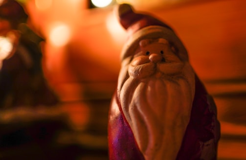 A small Santa Claus figurine with a long white beard and red attire stands out against a warm, blurred background. The lighting creates a cozy and festive atmosphere with soft highlights.