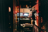 A dimly lit control room with multiple monitors displaying various screens. The room is cluttered with equipment, including a computer setup, control panels, and headsets. Shelves above the monitors hold miscellaneous items. Soft red and yellow lighting provides a warm ambiance.