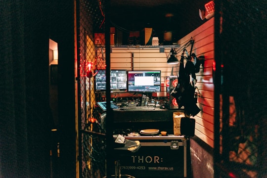 A dimly lit control room with multiple monitors displaying various screens. The room is cluttered with equipment, including a computer setup, control panels, and headsets. Shelves above the monitors hold miscellaneous items. Soft red and yellow lighting provides a warm ambiance.