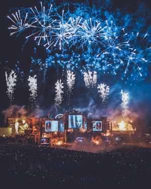 A vibrant night scene featuring a large outdoor festival or concert with a stage illuminated by lights and screens displaying artwork. Above the stage, a stunning display of blue and white fireworks lights up the sky. The area is filled with a dense crowd of people, contributing to an energetic atmosphere.