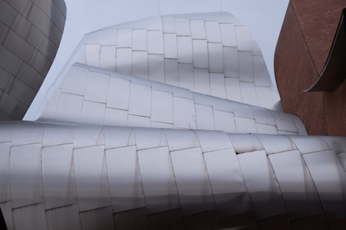 Architectural structure with sleek, metallic surfaces featuring curved and angular designs. The building exhibits a modern and abstract aesthetic with overlapping metal tiles and sections of brickwork.
