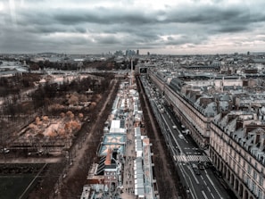 An aerial view of a European city featuring symmetrical streets, a bustling urban area with people walking along a market or fair, and vehicles on the road. On the left, a large park with bare trees and autumn foliage is visible. The architecture showcases classic European buildings with ornate rooftops. In the distance, a modern skyline with skyscrapers is seen under a cloudy sky.