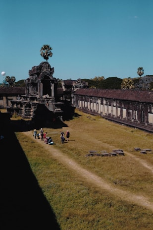 An ancient stone temple surrounded by grassy areas and palm trees. The architecture features intricate carvings and tall structures. A group of people, possibly tourists, walks along a path leading towards the temple.