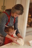 A woman and a small child are working together to knead dough on a lightly floured surface. The woman, wearing a red apron, is smiling and guiding the child's hands. The child, dressed in red, is focused on the dough. They are in a kitchen with soft, warm lighting.