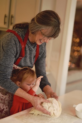 A woman and a small child are working together to knead dough on a lightly floured surface. The woman, wearing a red apron, is smiling and guiding the child's hands. The child, dressed in red, is focused on the dough. They are in a kitchen with soft, warm lighting.