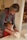 A woman and a small child are working together to knead dough on a lightly floured surface. The woman, wearing a red apron, is smiling and guiding the child's hands. The child, dressed in red, is focused on the dough. They are in a kitchen with soft, warm lighting.