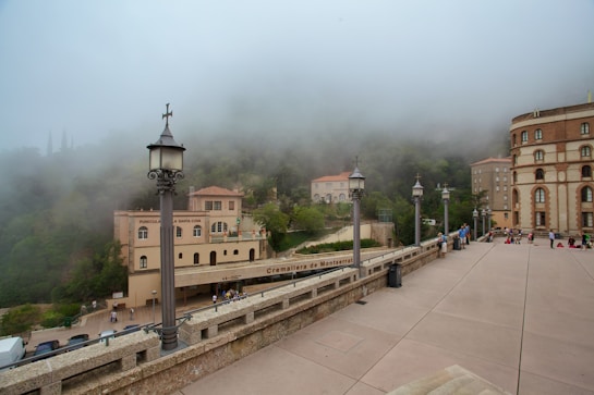 A misty scene of a mountainous area with historical architecture. The view includes multiple buildings nestled into lush greenery, partially obscured by fog. There are lamps lining a wide paved walkway, which is populated by a few people scattered about the area, and a structure labeled 'Cremallera de Montserrat' indicates a railway station or similar service.