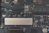 A close-up view of a computer motherboard, featuring various electronic components including capacitors, chips, and connectors. The design is intricate with a combination of black, silver, and copper colors visible on the circuit board.