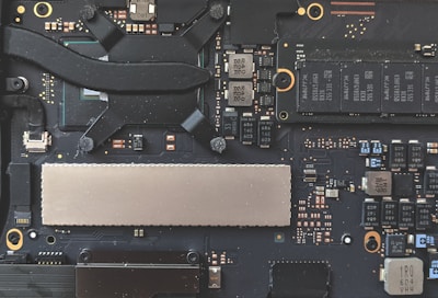 A close-up view of a computer motherboard, featuring various electronic components including capacitors, chips, and connectors. The design is intricate with a combination of black, silver, and copper colors visible on the circuit board.