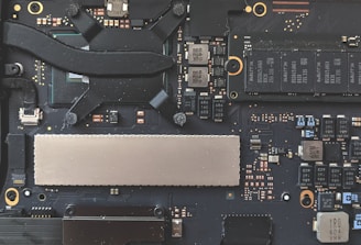 A close-up view of a computer motherboard, featuring various electronic components including capacitors, chips, and connectors. The design is intricate with a combination of black, silver, and copper colors visible on the circuit board.