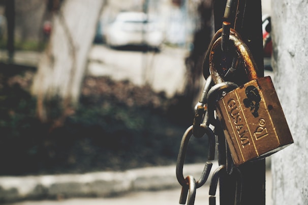 A close-up view of a brass padlock with a chain wrapped around it, attached to a metallic surface. The background is blurred, showing an outdoor setting with muted colors.
