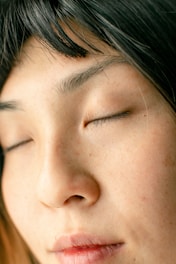 A close-up of a person's face with closed eyes, showcasing clear and smooth skin along with black hair framing the face. The image focuses on the serene and peaceful expression.