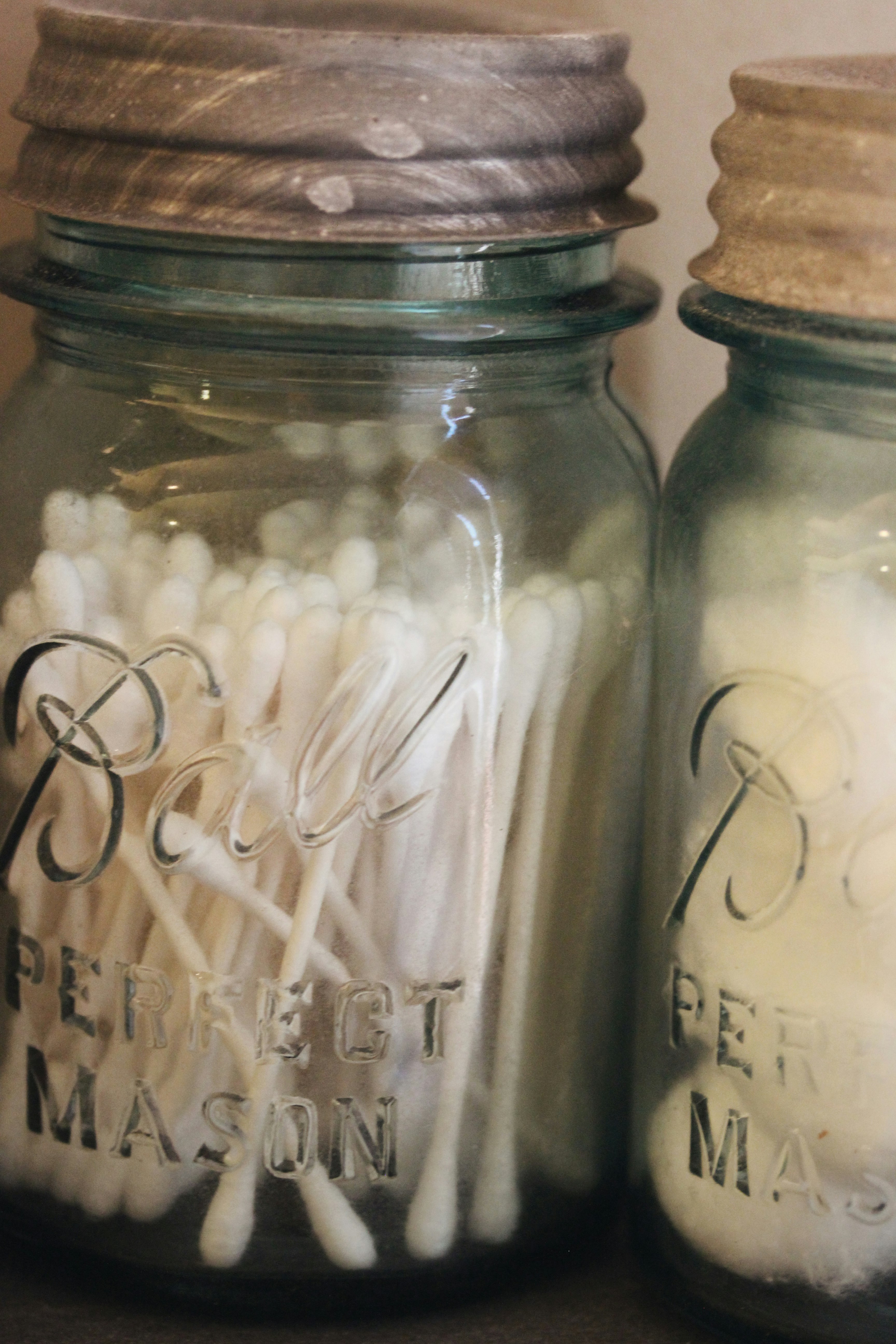 Bathroom essentials like Q-Tips and cotton swabs are stored in decorative vintage blue Ball glass canning jars, with logo and text writing imprinted on the sides and zinc silver lids on a shelf.