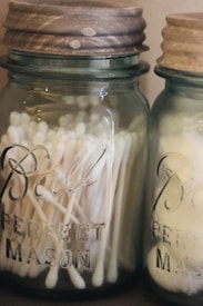 Two vintage mason jars with wooden lids are filled with cotton swabs and balls. The jars have a slightly tinted glass effect and the words 'Ball Perfect Mason' embossed on their surface.