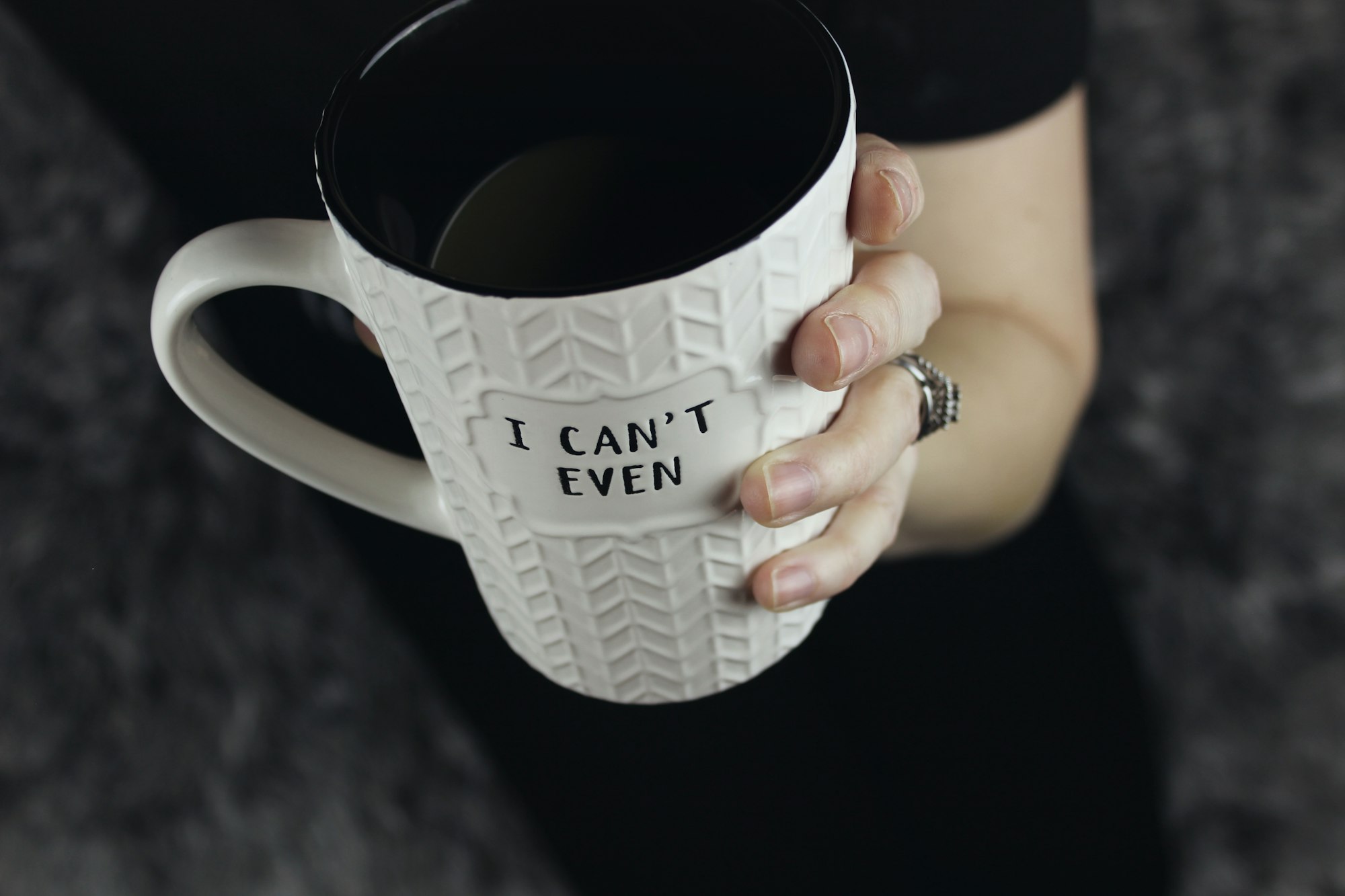 For those mornings when getting out of bed seems like the most difficult task in the world, this white coffee mug with herringbone pattern and texture tells it like it is: "I can't even..." held in a woman's hand with large wedding ring against a dark background.