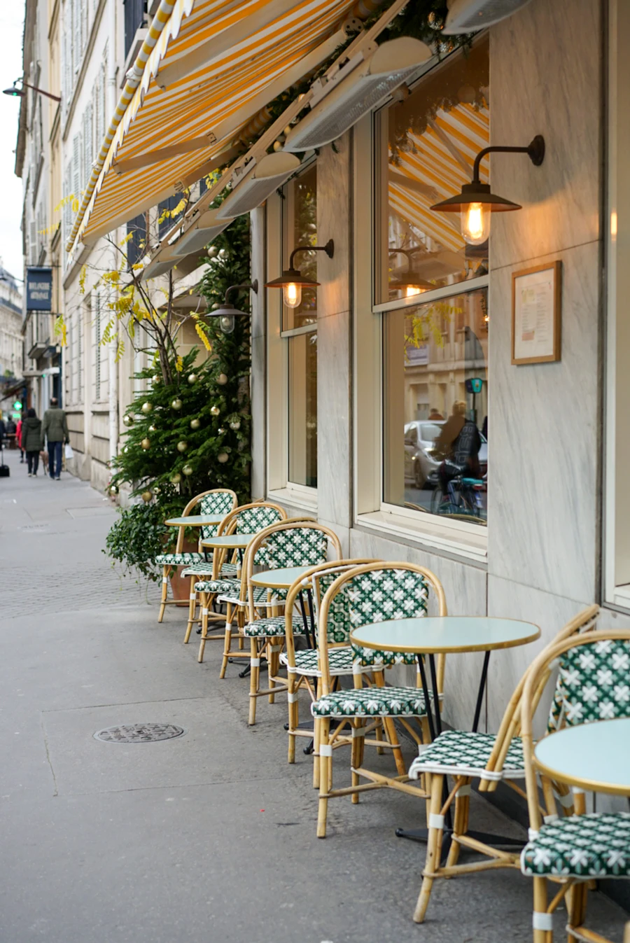Café tables and woven bistro chairs lined up outside under glowing wall lanterns and a striped awning