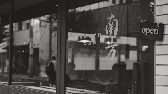A storefront with reflective glass showing the street outside, featuring an 'open' sign on the door and characters or text that may be decorative or informative. There is a knob and a lock on the door, and faint reflections of pedestrians and buildings can be seen.