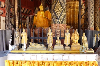A collection of golden Buddha statues is displayed on a decorative altar adorned with intricate patterns. The statues vary in posture, including seated, reclining, and standing forms. The backdrop features elaborately carved pillars and rich golden designs, adding to the overall ornate and sacred atmosphere.