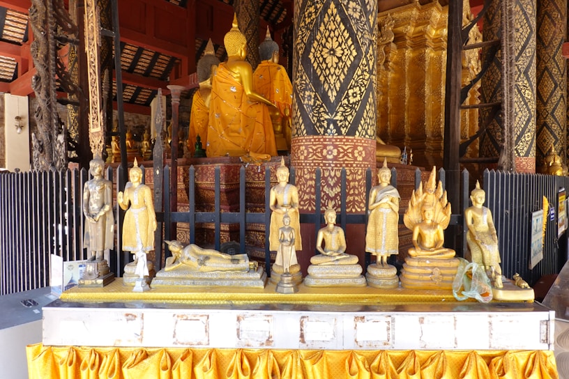 A collection of golden Buddha statues is displayed on a decorative altar adorned with intricate patterns. The statues vary in posture, including seated, reclining, and standing forms. The backdrop features elaborately carved pillars and rich golden designs, adding to the overall ornate and sacred atmosphere.