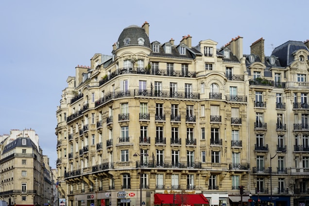 A large, elegant building with classic Parisian architecture characterized by beige stone facades and wrought-iron balconies. The building features multiple stories with numerous windows and a mansard roofline. Several shops are located on the ground floor, adding a vibrant touch to the scene.