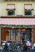 A storefront with wooden doors and large windows is adorned with decorative red and white striped awnings. Greenery and festive ornaments, including gold balls, decorate the windows and entrance. Three motorcycles and a cargo bike are parked in front of the establishment. The signage includes calligraphy in gold script and the address of the location.