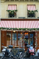 A storefront with wooden doors and large windows is adorned with decorative red and white striped awnings. Greenery and festive ornaments, including gold balls, decorate the windows and entrance. Three motorcycles and a cargo bike are parked in front of the establishment. The signage includes calligraphy in gold script and the address of the location.